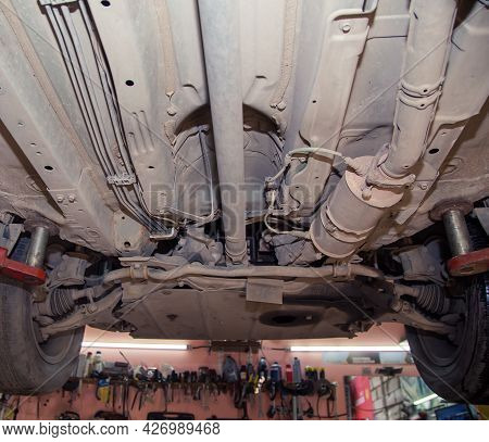 Bottom View Of The Underbody And Wheels Of An Old Car On A Lift. In The Garage, A Man Changes Parts 