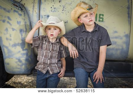 Two Young Boys Wearing Cowboy Hats Leaning Against an Antique Truck in a Rustic Country Setting. 
