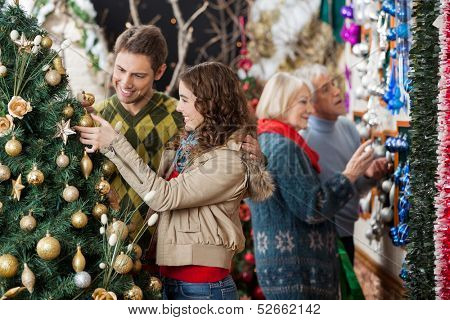 Happy young couple looking at Christmas tree with parents shopping in background at store