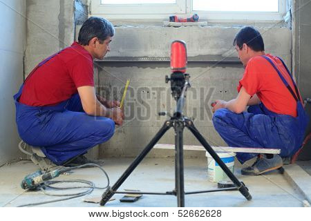 Two workers makes measurements with laser level tool in a niche for radiators in new apartment
