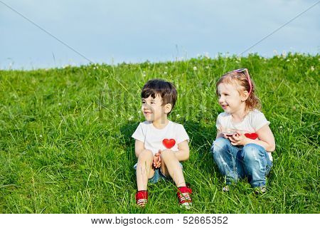 Two little children sit on edge of grassy slope, girl holds can lid with candies