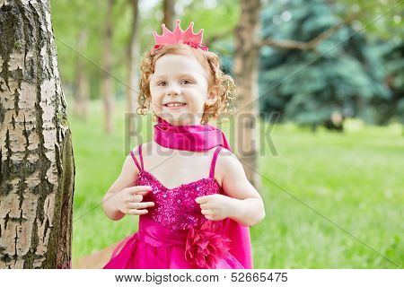 Half-length portrait of smiling little girl dressed in bright puffy gown who stands on grassy lawn in park 