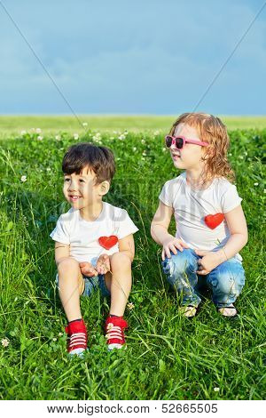 Two little children sit on edge of grassy slope, big green meadow behind them, red hearts pinned to theirs shirts