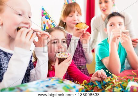 Children grabbing muffins at birthday party and cake, the kids are wearing hats, balloons and paper streamers for decoration 