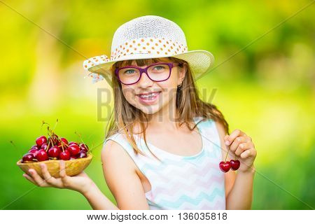 Child with cherries. Little girl with fresh cherries. Young cute caucasian blond girl wearing teeth braces and glasses. Portrait of a smiling young girl with bowl full of fresh cherries.