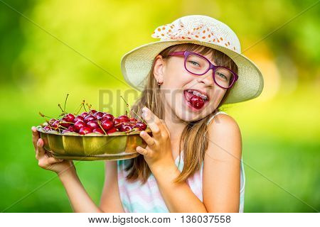 Child with cherries. Little girl with fresh cherries. Young cute caucasian blond girl wearing teeth braces and glasses. Portrait of a smiling young girl with bowl full of fresh cherries.