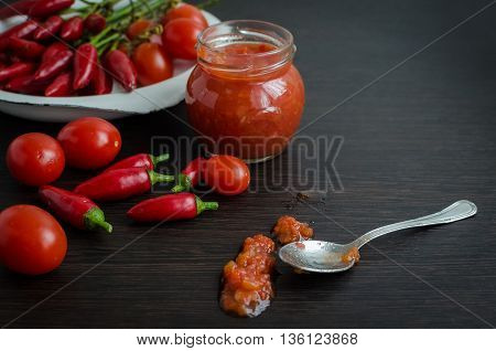 Home made ketchup in a jar. Tomato ketchup sauce with cherry tomatoes and mini red hot chili peppers in a small glass jar with a spoon on dark wooden background. Selective focus. Copy space.