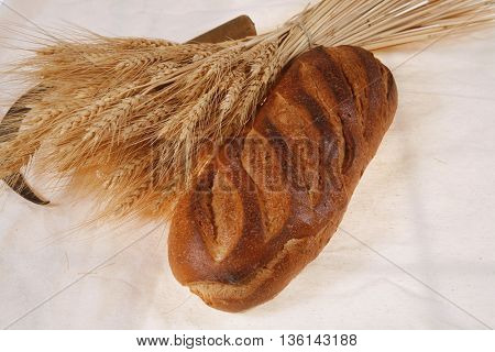 bunch of mown wheat ears with vintage handmade reaper hook sickle and rye dark white french fresh bread loaf on light linen tablecloth