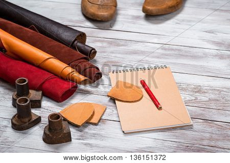 Brightly colored leather in rolls, working tools, shoe lasts, notebook with pencil on white wooden background. Leather craft.