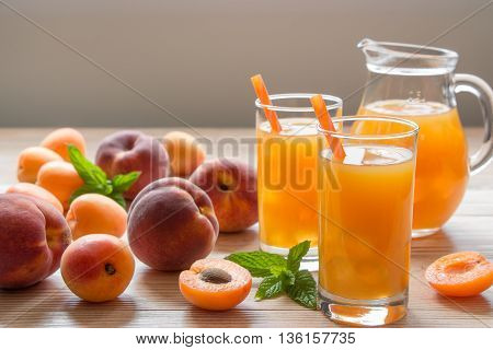 Apricot and peach juice in glasses with ice near the jar of juice and scattered apricots and peaches mint on a light wooden background. Apricot and peach juice with ice. Horizontal. Daylight. Close.