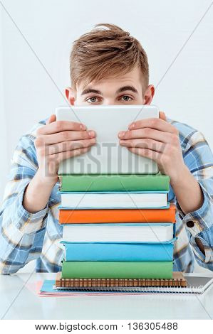 Modern problem: youth and computers. Young student using tablet computer while studying and looking at camera. Nice white interior with table full of books