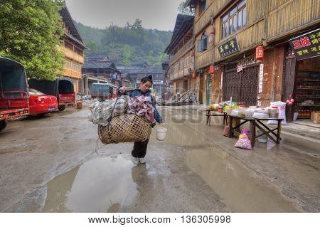 Zhaoxing Dong Village Guizhou China - April 8 2010: Street tradeswoman with a yoke on his shoulder carries a large wicker cage on street settlements of ethnic minorities passed wooden houses.