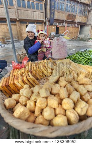 Zhaoxing Dong Village Guizhou Province China - April 8 2010: Street trade in the Asian countryside an elderly woman holding a child sells fried tofu.