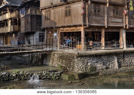 Zhaoxing Dong Village Guizhou Province China - April 8 2010: Chinese shopkeeper sits under a canopy of wooden huts near the cobblestone the embankment rural river.