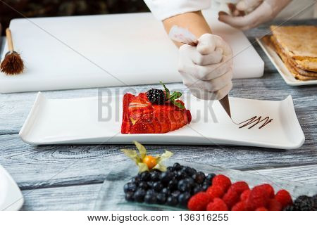 Blackberry and slices of strawberry. Hand with tool decorating dessert. Delicious strawberry flan. Liquid chocolate and berries.