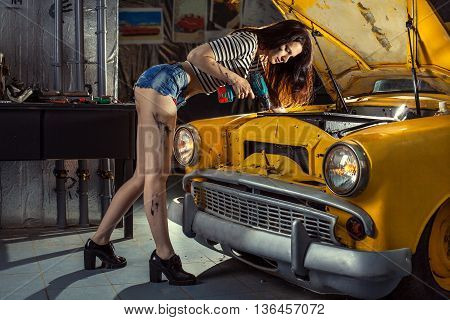 Woman mechanic in sexy shirts is repairing an old car with the screwdriver.