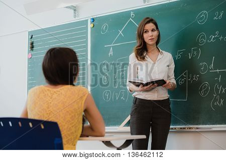 Teacher or docent or educator giving while lesson in front of a blackboard or board a sheet of paper and educate or teaching students or pupils or mates in a school or class.