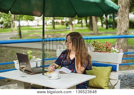 cute big size girl sitting and working on a summer ground in a cafe at a table under an umbrella and writes in a notebook near a laptop a cup and a glass of wine or water and cake.