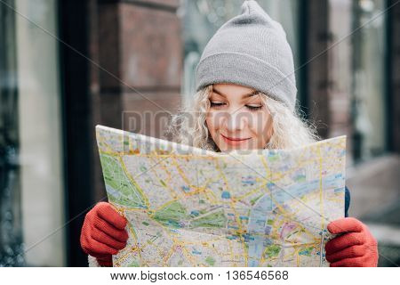 Young blond curly female tourist in warm hat and red gloves with London map looking for a way, winter