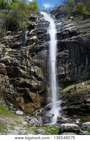 Narrow white stream of water flows over a stone cliff without touching the face of the rock until it splashes on the bedrock. The rock face is wetted by water running down the wall in several little trickles. Froda river, Chiggiogna, Swiss.