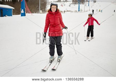Smiling woman and girl in red jackets on snowy slope at ski resort.