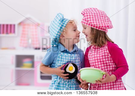 Little girl and boy in chef hat and apron cooking fried eggs in toy kitchen. Wooden toys for young children. Kids play and cook at home or daycare. Toddler kid playing with stove pans and dishes.