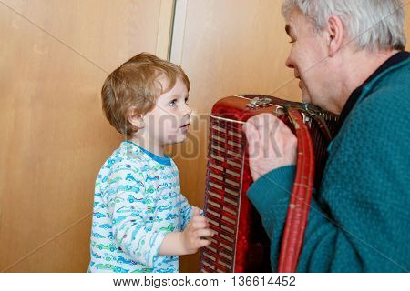 Happy blond little kid boy and his grandfather playing together with accordion. Senior man teaching his grandson, cute toddler to play with music instrument at home.