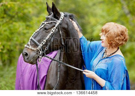 Red-haired smiling woman in blue capote with bay horse in the park.