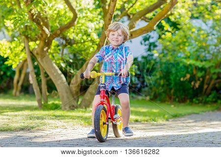 funny blond kid boy in colorful clothes driving bicycle in domestic garden. Toddler child dreaming and having fun on warm summer day. Active games for children outdoors.
