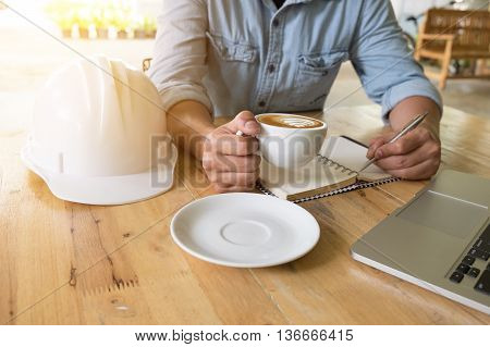 engineering useing laptop computer note someting with pen at notbook and holding a cup of coffee on wooden table