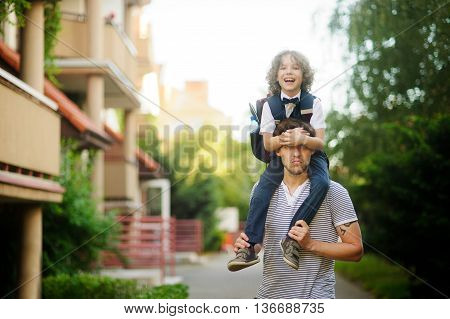 Little schoolboy sitting on the shoulders of his father. The father holds the boy's legs. The boy delighted. He likes to joke with dad. Son happily smiling and father jokingly serious.