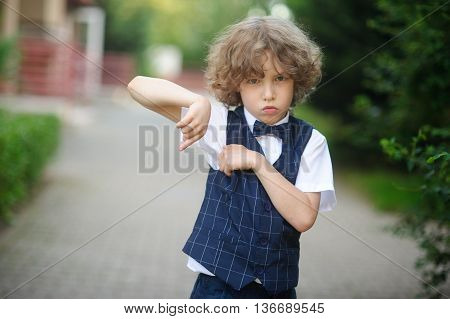 Little pupil is in the schoolyard with an angry expression on his face. The boy has blond curly hair and blue eyes. He is angry and showing something with his hands.
