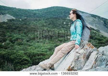 Beautiful traveler young woman sitting on peak of cliff and enjoying view of mountains in summer outdoor