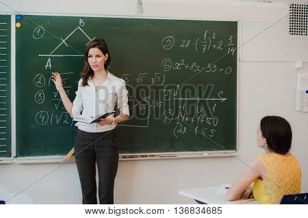 Teacher or docent or educator giving while lesson in front of a blackboard or board a sheet of paper and educate or teaching students or pupils or mates in a school or class.