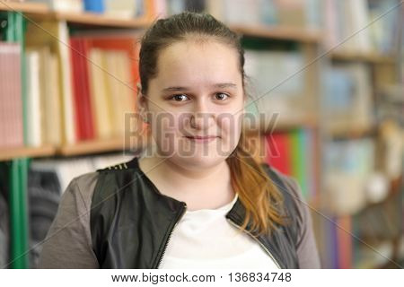 half growth portrait of student with hair gathered in ponytail on background of shelves with books, shyly smiling at camera