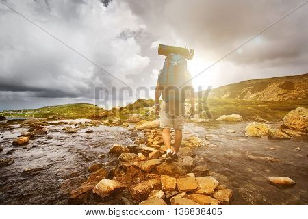 Hiker man with backpack crossing a river on stones in bulgarian mountains. Hiking and leisure theme. Image with sunlight effect