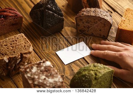 Hand takes blank business card of professional artisan baker presented in center of many mixed alternative baked exotic bread samples above wooden rustic table