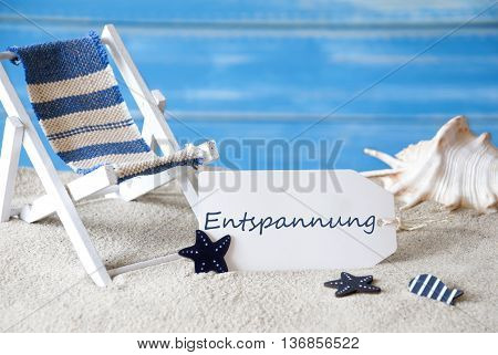 Summer Label With German Text Entspannung Means Relaxation. Blue Wooden Background. Card With Holiday Greetings. Beach Vacation Symbolized By Sand, Deck Chair And Shell.