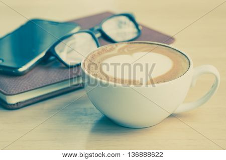 A cup of coffe with heart pattern in a white cup on smartphone leather notebook and eyeglasses on wooden table.Close up.Vintage tone