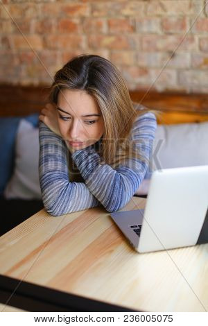 Young Woman Sitting In Cozy Coffee House Near Wooden Table With Glass Cup Of Cappuccino And Using La