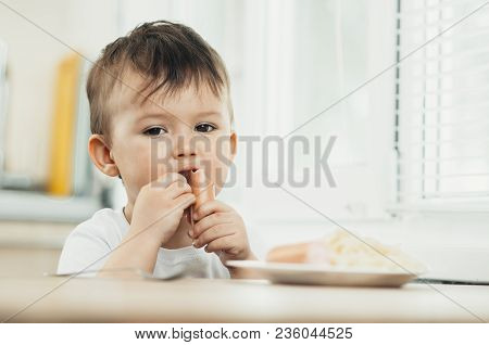 The Child Is Sitting In The Kitchen And Greedily Eats Sausage