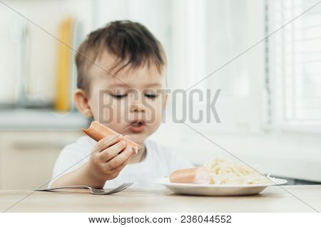 The Child Is Sitting In The Kitchen And Greedily Eats Sausage