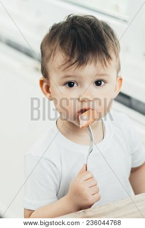 The Child Is Sitting In The Kitchen And Greedily Eats Sausage