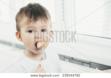 The Child Is Sitting In The Kitchen And Greedily Eats Sausage