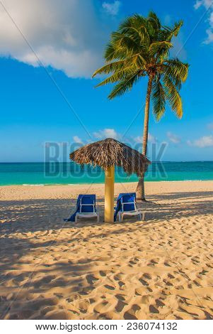 Holguin, Cuba, Playa Esmeralda. Umbrella And Two Lounge Chairs Around Palm Trees. Tropical Beach On 