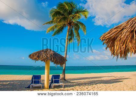 Holguin, Cuba, Playa Esmeralda. Umbrella And Two Lounge Chairs Around Palm Trees. Tropical Beach On 