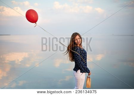 Portrait Of Young Woman With Red Air Balloon And Present Bag Near The Calm Sea Or Lake. Clouds Are R