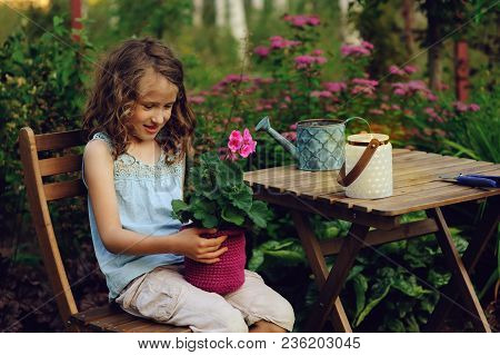 Dreamy Happy Child Girl Relaxing In Summer Evening Garden With Heranium Flower In Pot. Teaching Kids