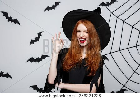 Halloween Witch Concept - Happy Halloween Ginger Hair Witch Holding Ok Sign With Fingers Posing Over