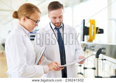 Bearded Technologist And His Pretty Colleague Wearing White Coats Standing At Modern Equipment Of Da